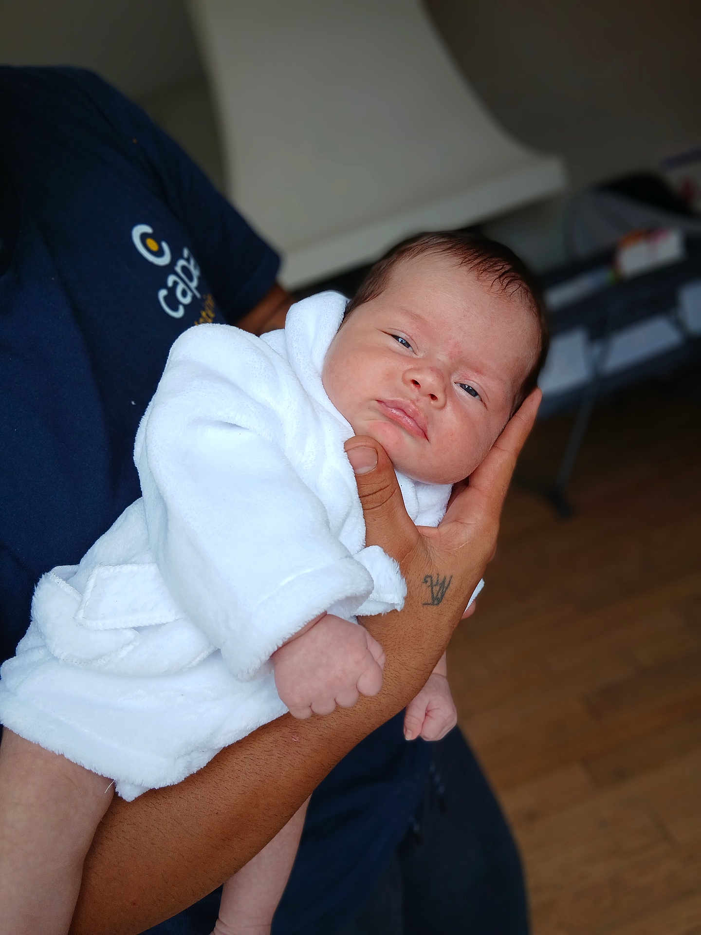 Marta a rejoint le concours — aidez-le/la à gagner de superbes lots ! baby, infant, white_robe, hand, tattoo, person, indoor, wooden_floor, blurred_background, cute, sleepy, skin, holding, portrait, newborn, clothing, human, face, expression, close_up