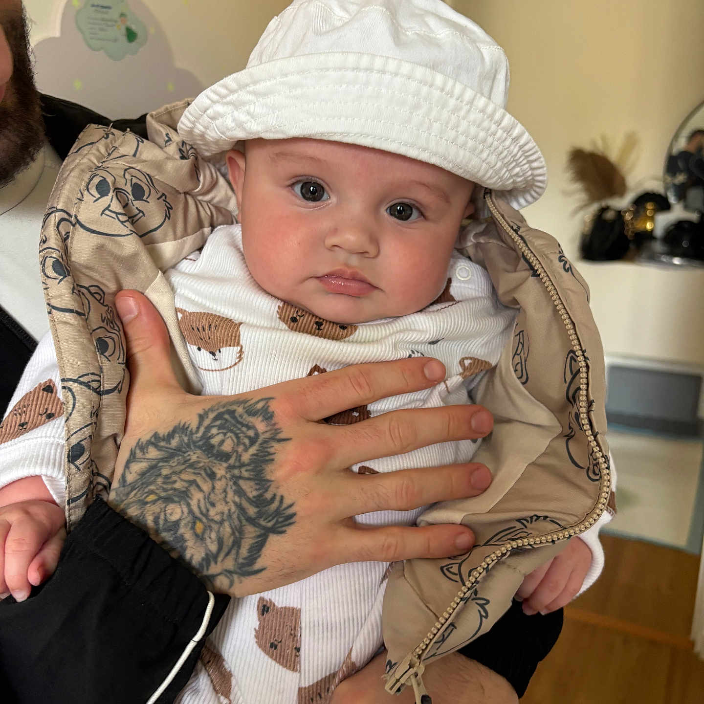 Aylann participe au concours pour gagner de l'argent avec cette photo : adult_hand, animal_print, baby, big_eyes, bucket_hat, child, chubby_cheeks, closeup, holding, indoor, infant, jacket, mirror_reflection, nursery_decor, onesie, portrait, tattoo, white_hat, wooden_floor, zipper