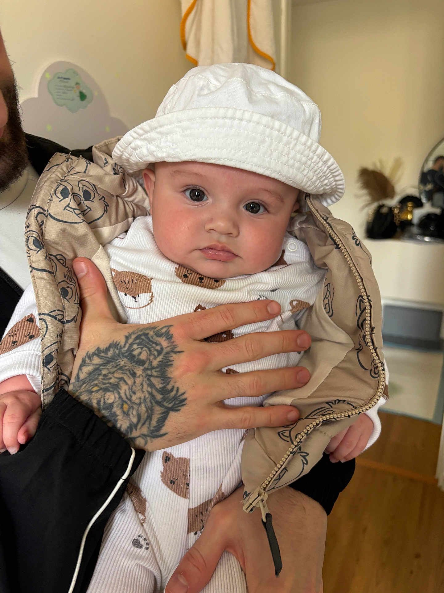 Aylann participe au concours pour gagner de l'argent avec cette photo : baby, infant, child, bucket_hat, white_hat, onesie, animal_print, tattoo, adult_hand, jacket, zipper, big_eyes, chubby_cheeks, portrait, indoor, mirror_reflection, nursery_decor, wooden_floor, holding, closeup