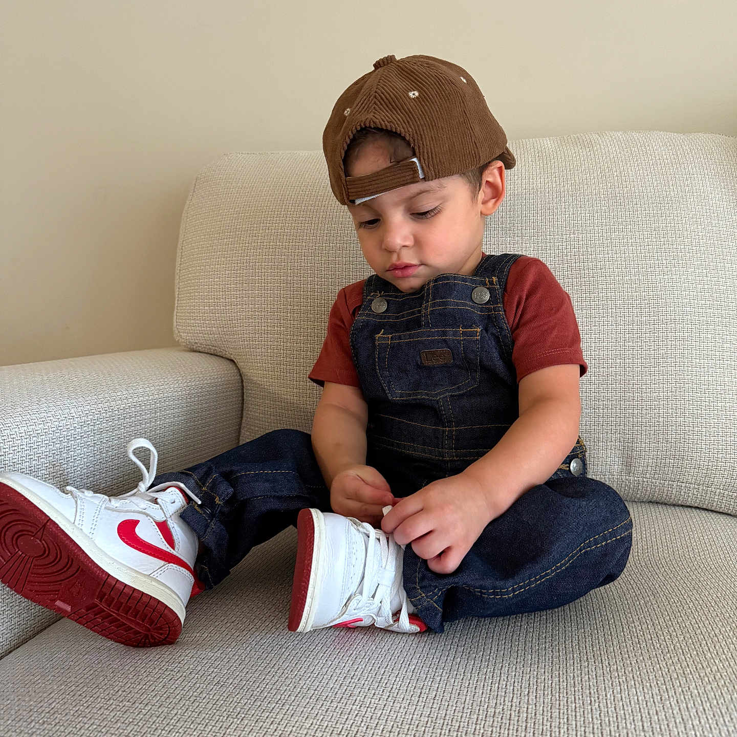 Maximiliano joined the competition — help win amazing prizes! beige_couch, brown_cap, cap, casual_clothing, child, couch, cute, denim_overalls, focused, footwear, hands, indoor, red_shirt, red_sneakers, seated, shoe_laces, sneakers, toddler, white_sneakers, young_child