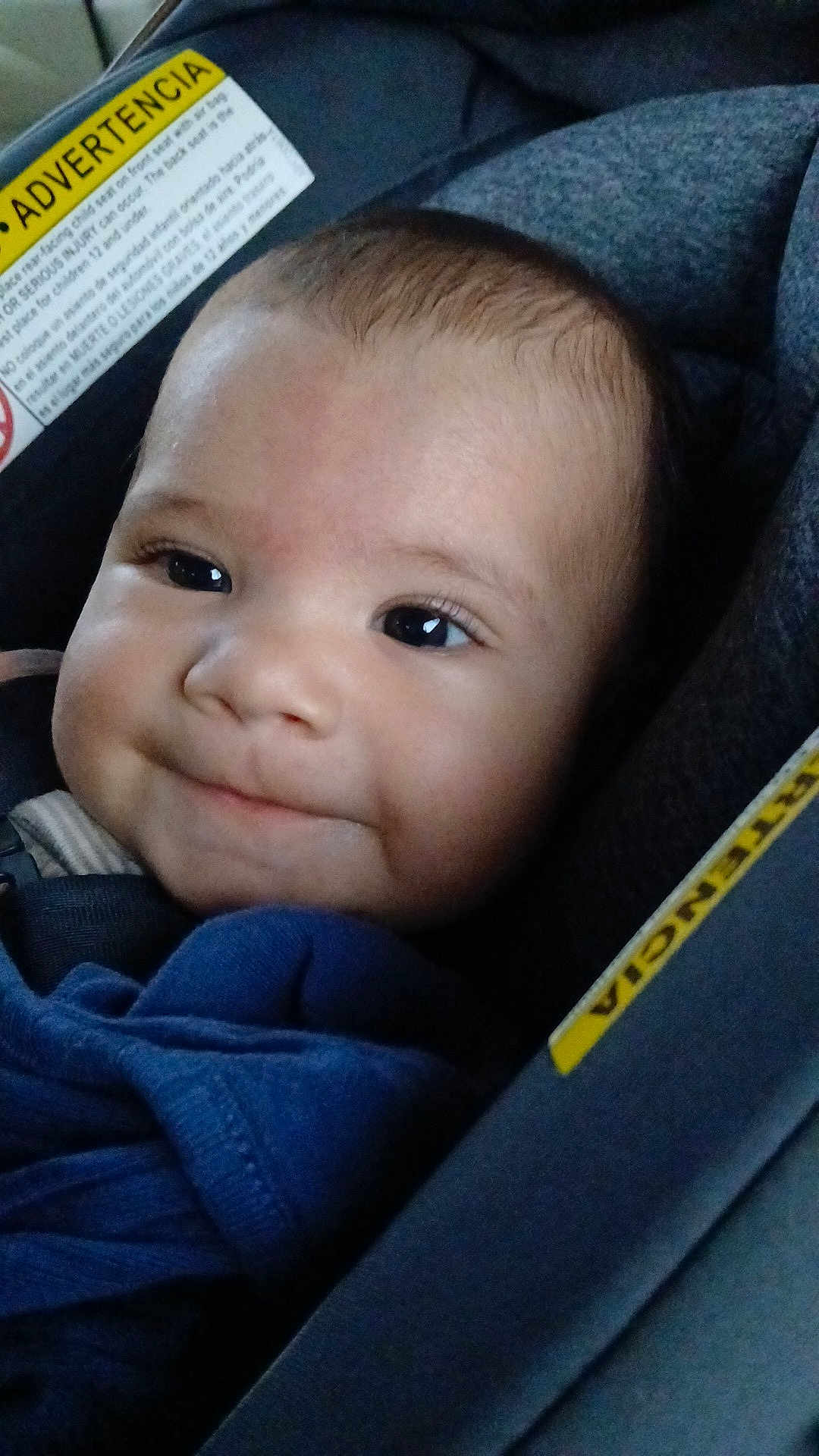 Raphael is registered to the contest to win money with this photo: baby, blue_blanket, car_seat, child, close_up, clothing, cute, eyelashes, face, happy, head, indoors, infant, person, safety_label, seat, seat_belt, skin, smile, young_child