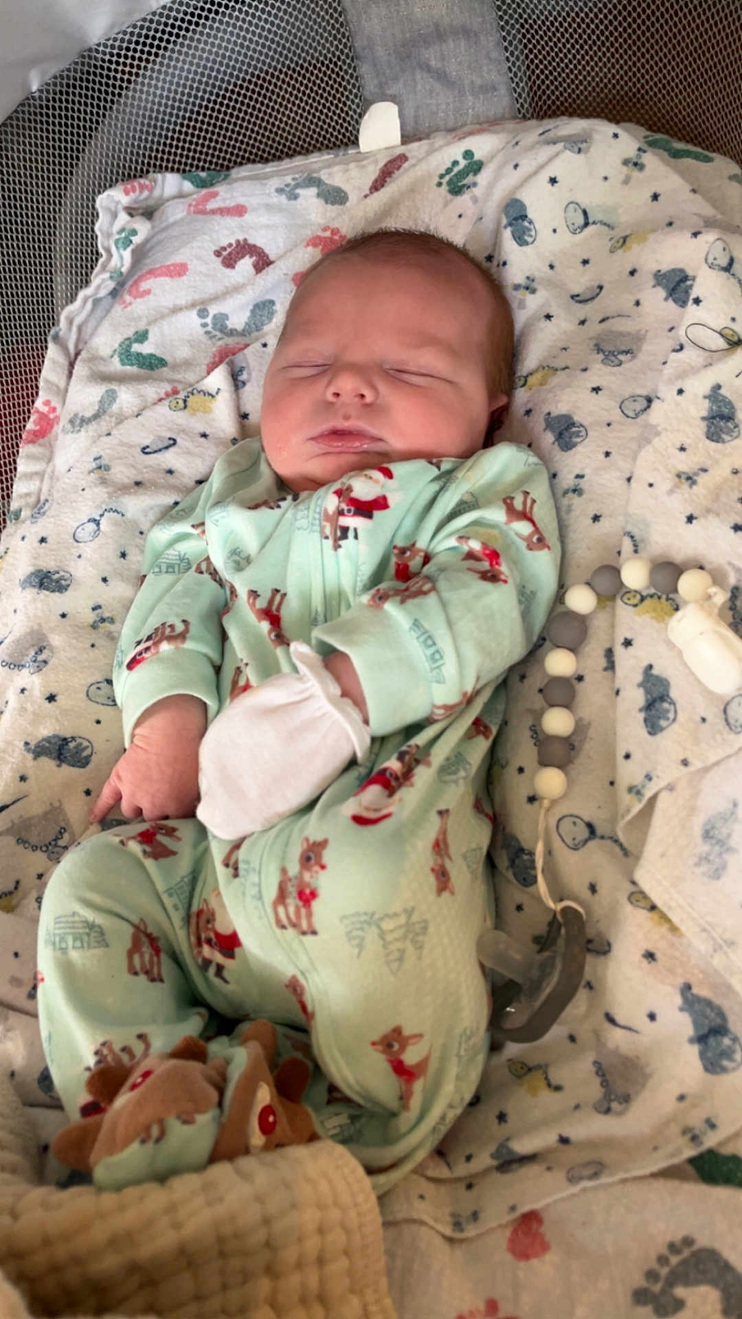 Gunnison is registered to the contest to win money with this photo: newborn, baby, sleeping, onesie, reindeer, blanket, crib, toy, mittens, pacifier, soft_toy, footprint_pattern, baby_clothing, infant, cozy, indoors, cute, resting, child, peaceful