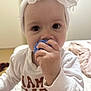 baby, child, headband, white_clothing, indoor, toy, hand, face, closeup, cute, infant, young_child, portrait, bed, soft_lighting, human, expression, sitting, curious, bow