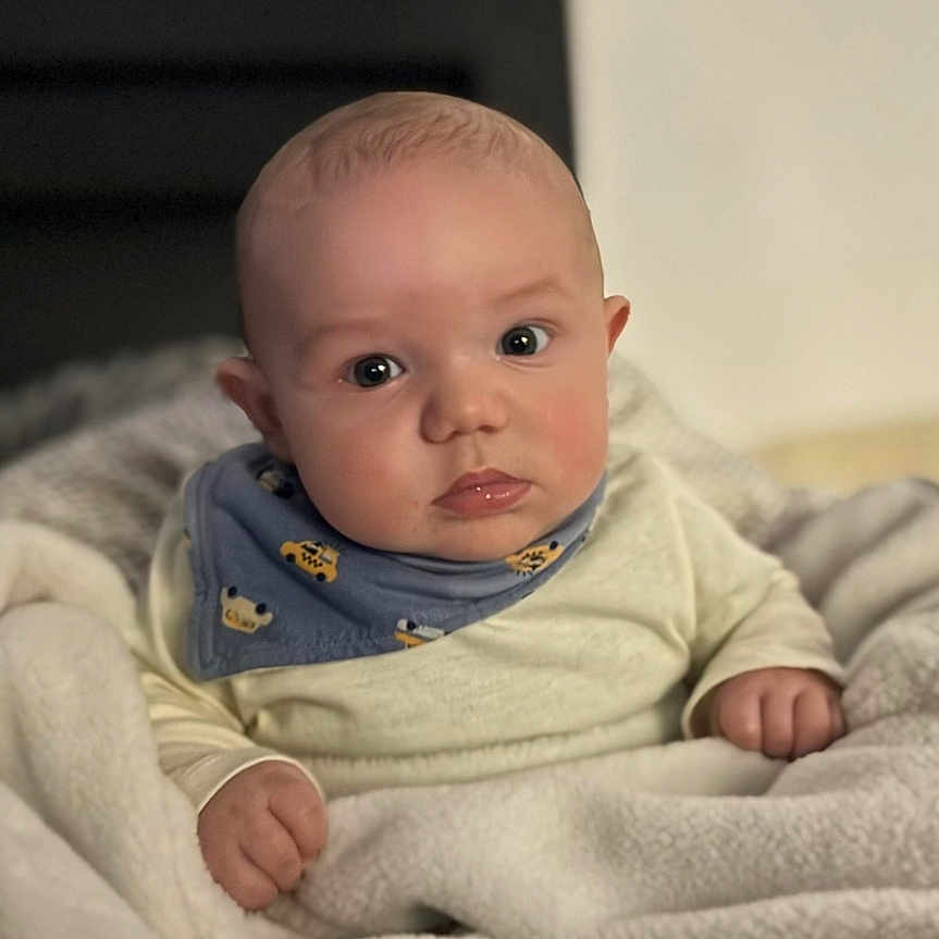 Kenny participe au concours pour gagner de l'argent avec cette photo : baby, blanket, blue_bib, child, chubby_cheeks, clothing, curious, cute, face, hands, head, indoors, infant, person, portrait, sitting, soft_texture, warm, wide_eyes, young