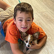Papi is registered to the contest to win money with this photo: child, boy, dog, puppy, indoor, floor, orange_shirt, light_wood_floor, smiling, hugging, pet, cute, young, ears, casual_clothing, happy, playing, domestic_animal, leash, close_up