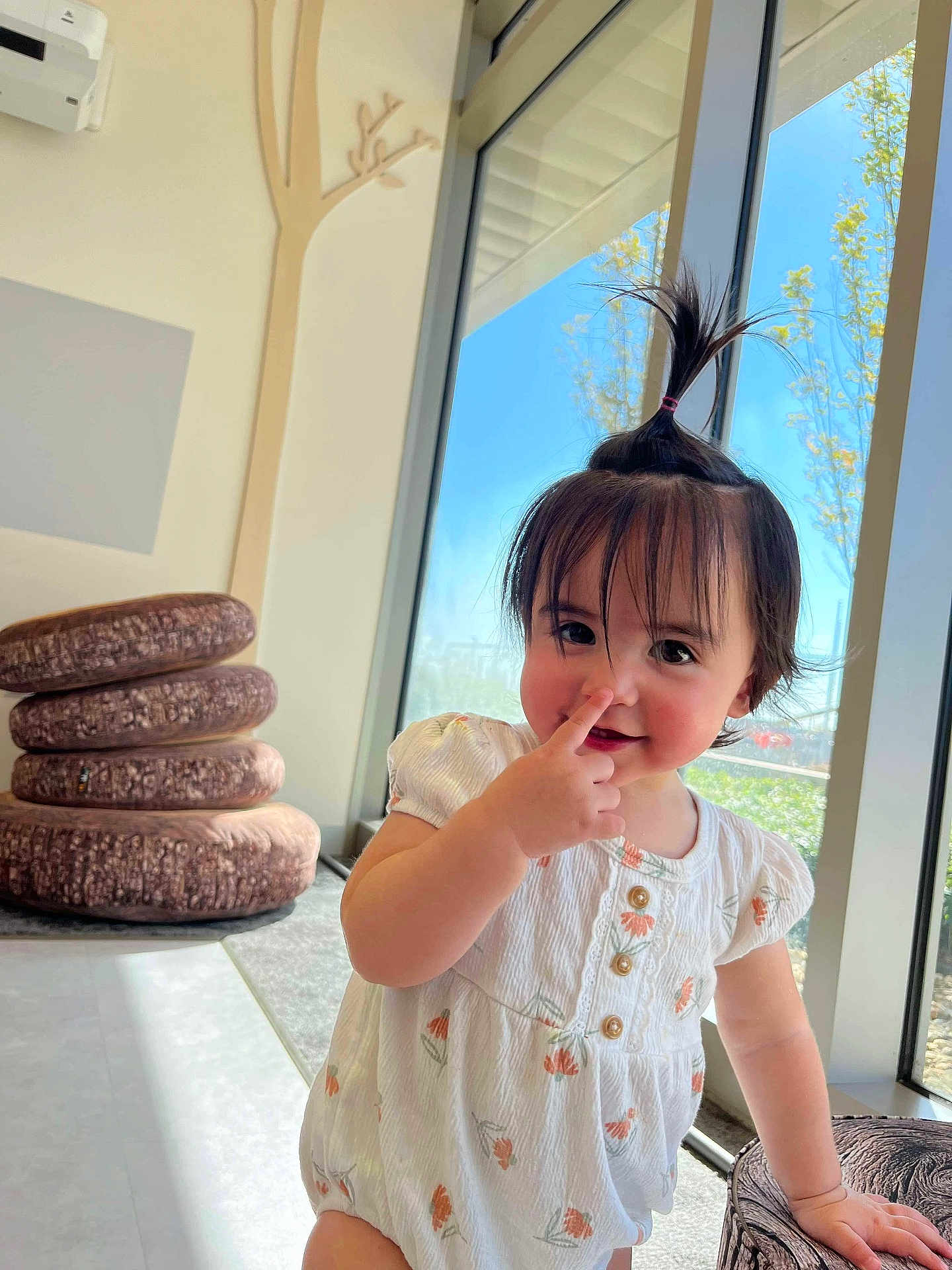 Alice is registered to the contest to win money with this photo: toddler, child, cute, indoors, window, sunlight, hair_ponytail, playful, nose_picking, white_outfit, floral_pattern, cushions, stacked_cushions, floor, hand, smile, face, person, young_child, bright