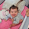 baby, smiling, child, plush_toy, cow_plush, pink_blanket, pillow, playpen, tattooed_hand, cute, indoors, happy, infant, clothing, sitting, person, soft_toy, cozy, face, hair