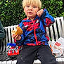 toddler, child, blonde_hair, blue_eyes, spiderman_jacket, black_pants, bench, burger, food, crumbs, toy, fire_truck, greenery, outdoor, casual_clothing, messy, eating, portrait, daylight, person