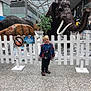child, boy, indoor, museum, display, woolly_mammoth, prehistoric_animals, white_fence, smiling, blue_jacket, black_pants, shoes, exhibit, shopping_mall, sign, modern_architecture, glass_roof, floor_tiles, bag, curious