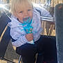 child, toddler, blonde_hair, blue_ice_cream, messy, white_hoodie, black_pants, black_shoes, red_swoosh, metal_chair, outdoor, sunlight, happy, face_paint, ice_cream_cone, messy_face, playful, sitting, food, summer
