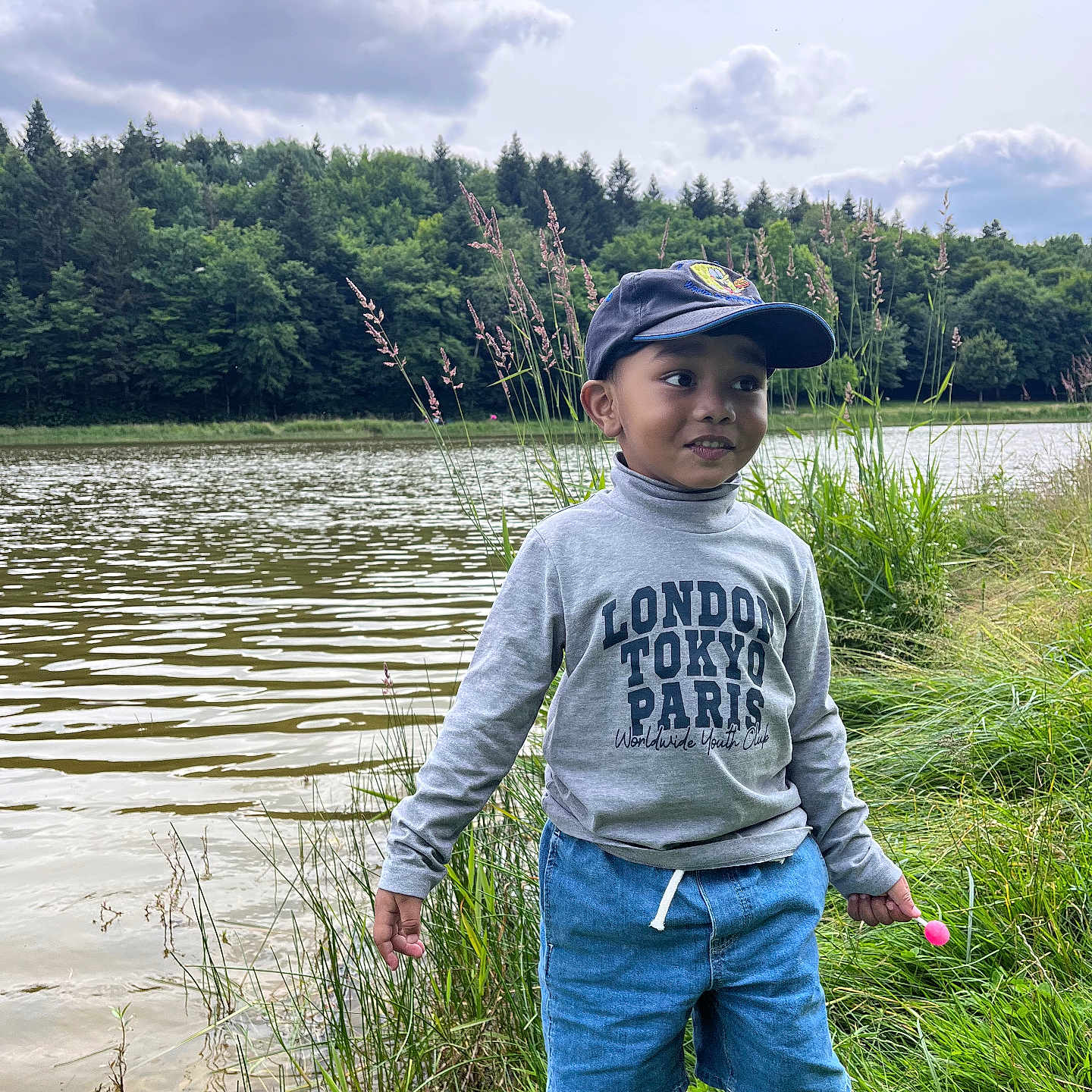 Zaki participe au concours pour gagner de l'argent avec cette photo : boy, cap, casual_clothing, child, clouds, daytime, grass, greenery, lake, nature, outdoor, person, portrait, scenery, shorts, standing, summer, sweatshirt, trees, water