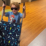Bryson is registered to the contest to win money with this photo: child, toddler, sunglasses, stroller, indoor, wood_floor, dinosaur_pattern, blue_shirt, curly_hair, toy, play, person, flooring, wall, door, hand, footwear, adult, fun, pretend_play