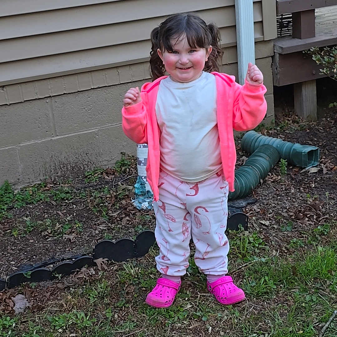 Kinsley is registered to the contest to win money with this photo: child, girl, pigtails, pink_jacket, pink_shoes, white_shirt, pants, outdoor, grass, yard, house_siding, garden_hose, smiling, standing, casual_clothing, playful, person, daylight, nature, happy