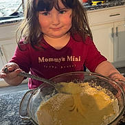 Kinsley is registered to the contest to win money with this photo: child, girl, kitchen, mixing_bowl, batter, spoon, countertop, cabinet, red_shirt, long_hair, smile, heart_doodle, indoor, cooking, playful, young, appliance, home, person, food_preparation