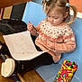 toddler, child, drawing, marker, paper, pigtails, sweater, black_pants, bench, wooden_bench, colorful_container, markers, indoor, focused, art, creative, girl, seat, clothing, hair_ties