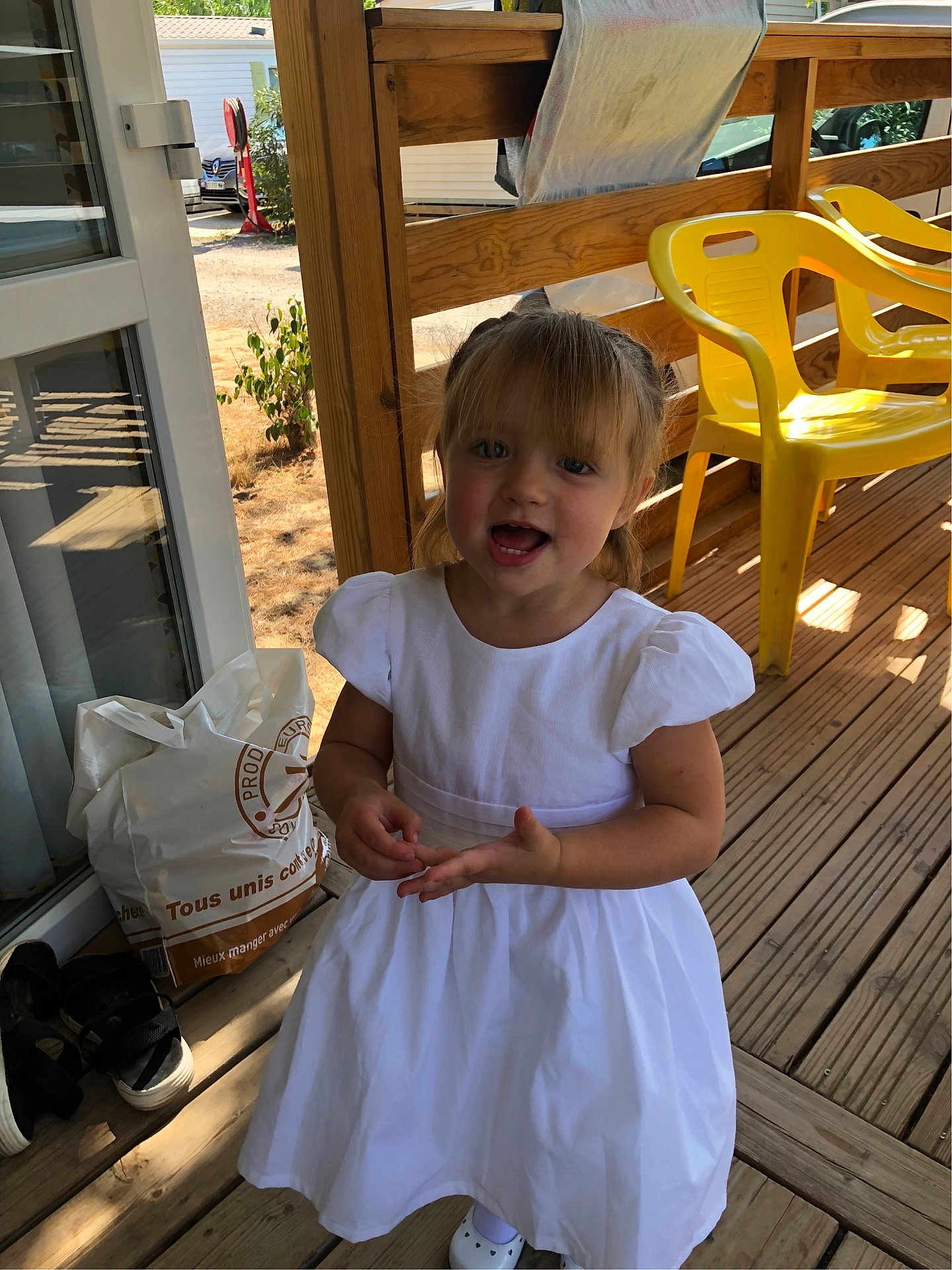 Chloé a rejoint le concours — aidez-le/la à gagner de superbes lots ! child, girl, white_dress, smiling, outdoor, wooden_deck, yellow_chair, sunlight, shadow, railing, bag, shoes, casual, happy, person, daylight, porch, expression, cute, standing