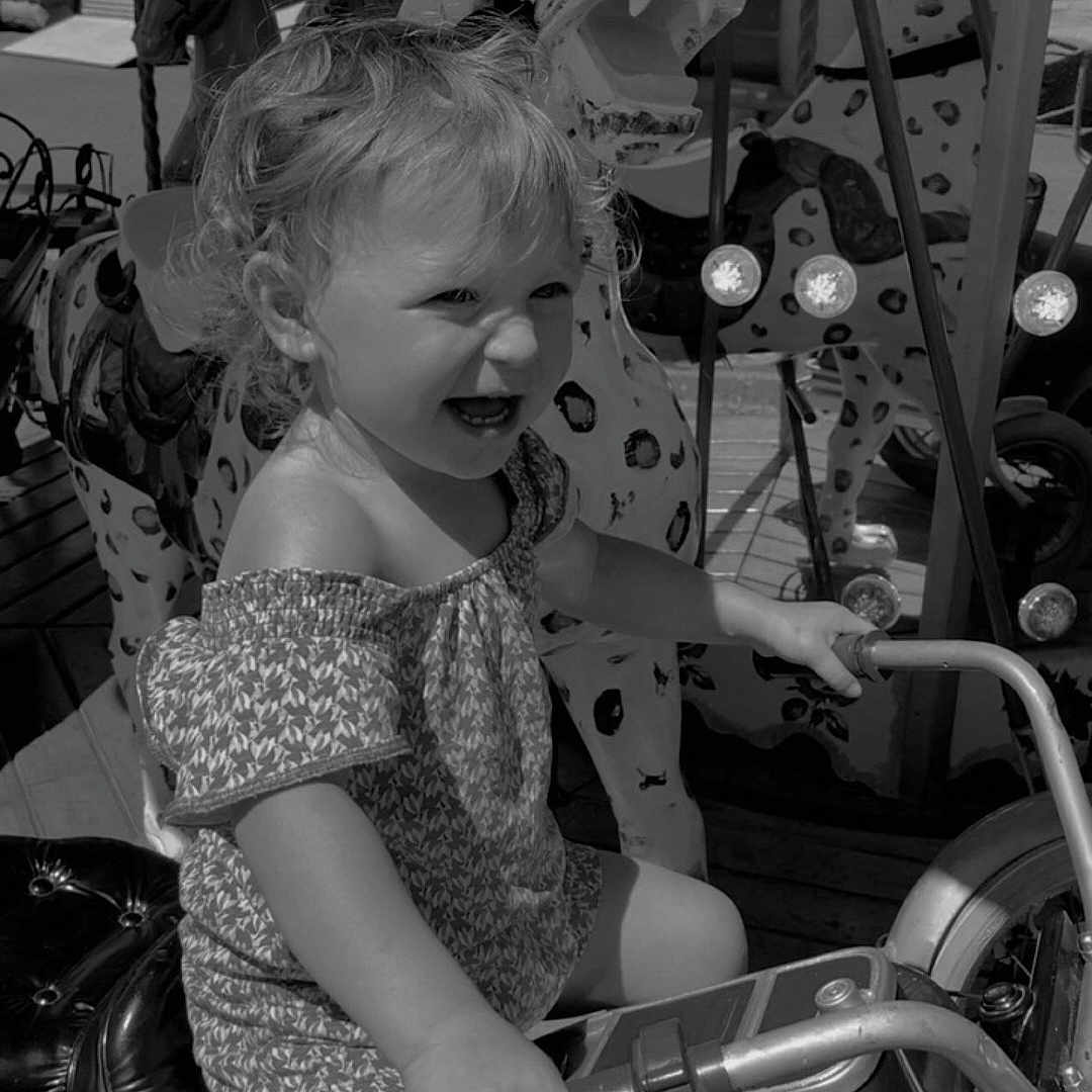 Paolina participe au concours pour gagner de l'argent avec cette photo : amusement_park, animal_figure, black_and_white, carousel, child, clothing, daylight, fun, hair, handlebar, happy, motorcycle, outdoor, playground, riding, seat, shorts, smiling, toddler, toy