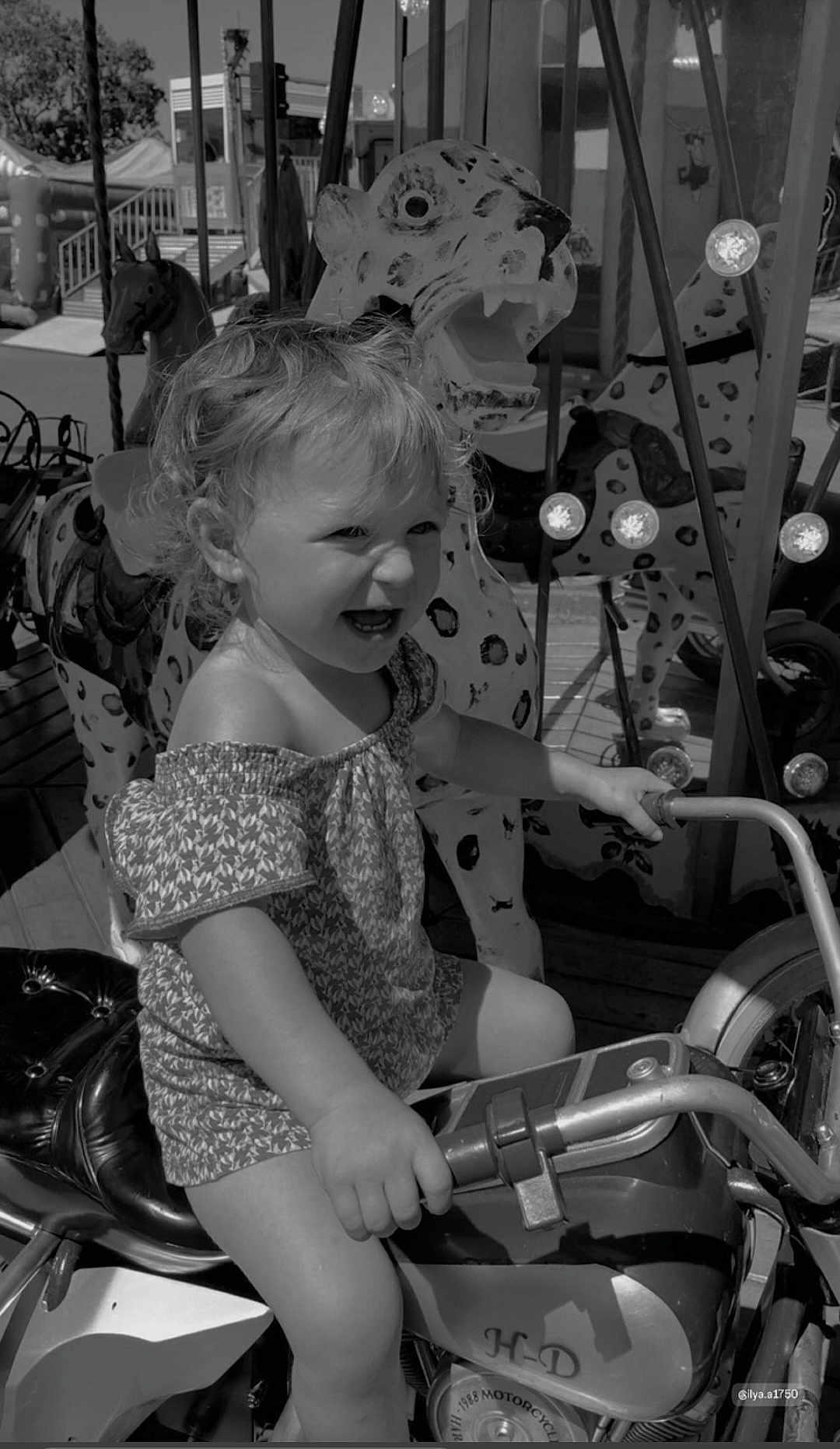 Paolina participe au concours pour gagner de l'argent avec cette photo : child, toddler, motorcycle, carousel, animal_figure, playground, outdoor, smiling, happy, riding, toy, seat, handlebar, clothing, shorts, hair, fun, daylight, amusement_park, black_and_white