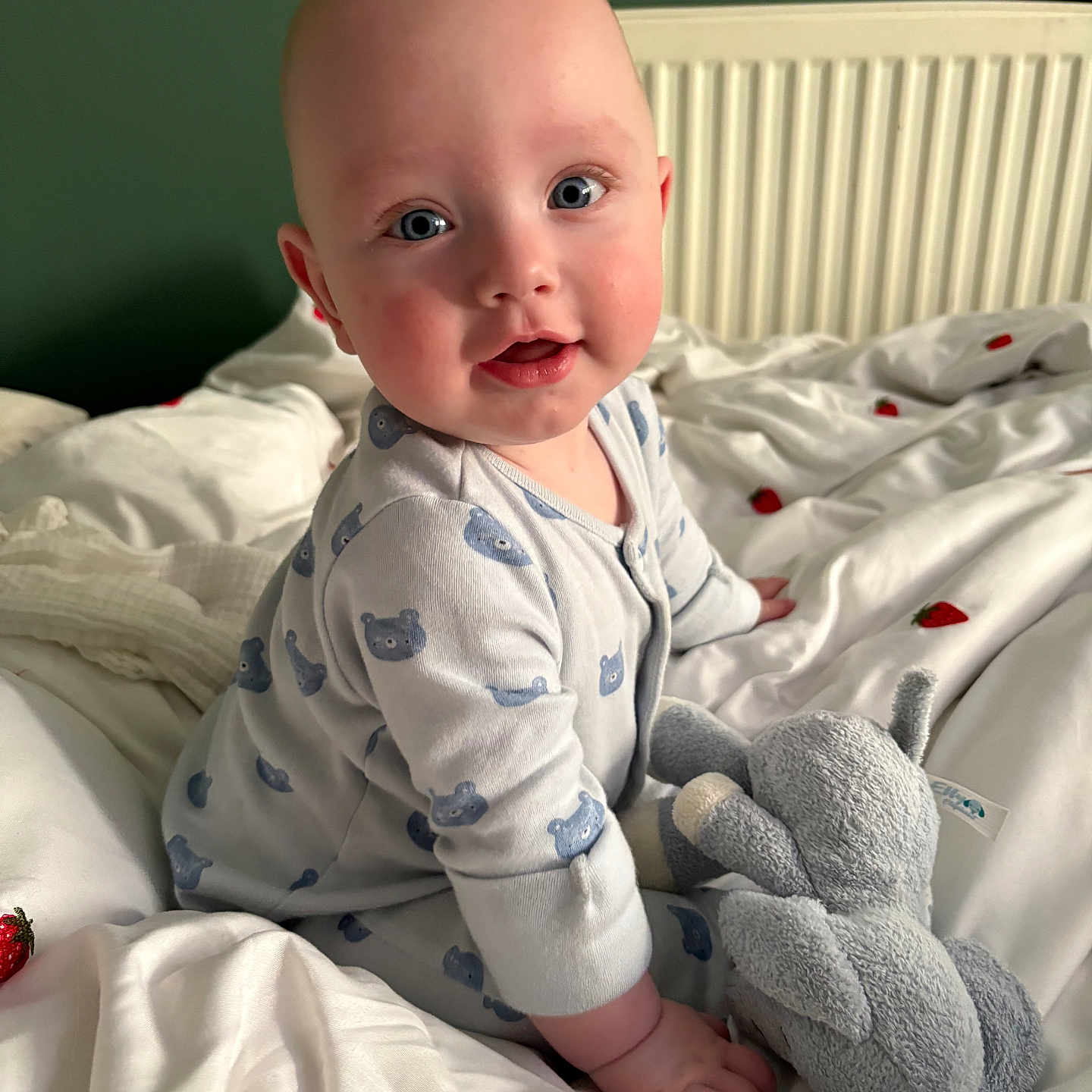 Hugo is registered to the contest to win money with this photo: baby, bed, bedspread, blue_eyes, child, cute, elephant_toy, face, green_wall, hand, indoor, infant, onesie, person, plush_toy, radiator, sitting, smiling, strawberry_pattern, white_bed