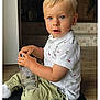 toddler, child, boy, indoor, sitting, blue_eyes, white_shirt, polo_shirt, green_pants, holding, bottle, floor, wooden_floor, mat, curious, casual_clothing, portrait, person, young_child, cute