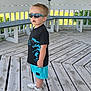 bench, boy, casual_clothing, child, daylight, dinosaur_graphic, gazebo, greenery, outdoor, shade, shorts, side_view, sneakers, standing, summer, sunglasses, trees, tshirt, wooden_deck, young_child