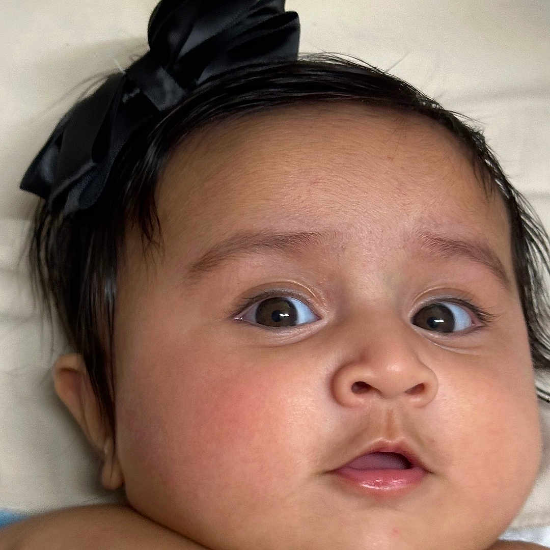 Aanvi joined the competition — help win amazing prizes! adorable, baby, black_bow, cheeks, child, closeup, curious, cute, expression, face, head, indoors, infant, lying_down, mouth, person, portrait, skin, soft_surface, wide_eyes