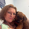 woman, glasses, tattoos, puppy, dog, pet, indoor, smile, person, holding, brown_dog, cute, affection, face, portrait, casual_clothing, light_blue_wall, sign, friendly, animal