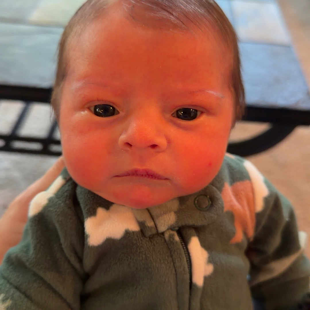 Emerson is registered to the contest to win money with this photo: baby, child, infant, portrait, sitting, face, eyes, serious, indoors, onesie, clothing, patterned_clothing, green_clothing, zipper, head, hair, fair_skin, table, furniture, close_up