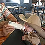baby, cowboy_hat, bottle, feeding, adult_hand, table, chair, cafe, indoor, person, milk, clothing, wood_floor, bib, wood_table, hat, child, face, seated, window