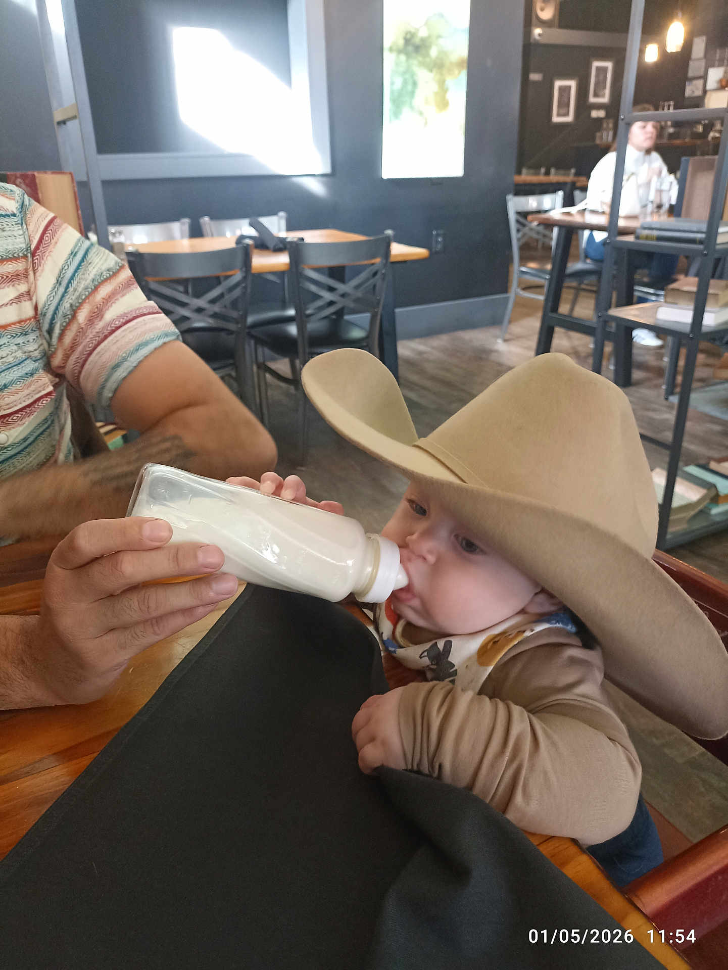 Axel joined the competition — help win amazing prizes! baby, cowboy_hat, bottle, feeding, adult_hand, table, chair, cafe, indoor, person, milk, clothing, wood_floor, bib, wood_table, hat, child, face, seated, window