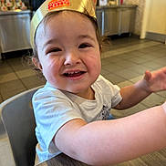 Luka is registered to the contest to win money with this photo: toddler, child, smile, burger_king, crown, fast_food, restaurant, table, white_shirt, happy, face, curly_hair, indoor, high_chair, arm, expression, food_court, background, person, closeup