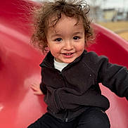 Luka joined the competition — help win amazing prizes! child, toddler, curly_hair, smiling, slide, playground, outdoor, black_jacket, black_pants, red_slide, happy, person, young_child, fun, park, casual_clothing, cute, portrait, daylight, active
