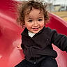 child, toddler, curly_hair, smiling, slide, playground, outdoor, black_jacket, black_pants, red_slide, happy, person, young_child, fun, park, casual_clothing, cute, portrait, daylight, active