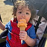 child, toddler, ice_cream, cone, curly_hair, spiderman_shirt, stroller, sunlight, outdoor, summer, snack, cute, food, happy, portrait, baby, person, seatbelt, casual_clothing, sweet