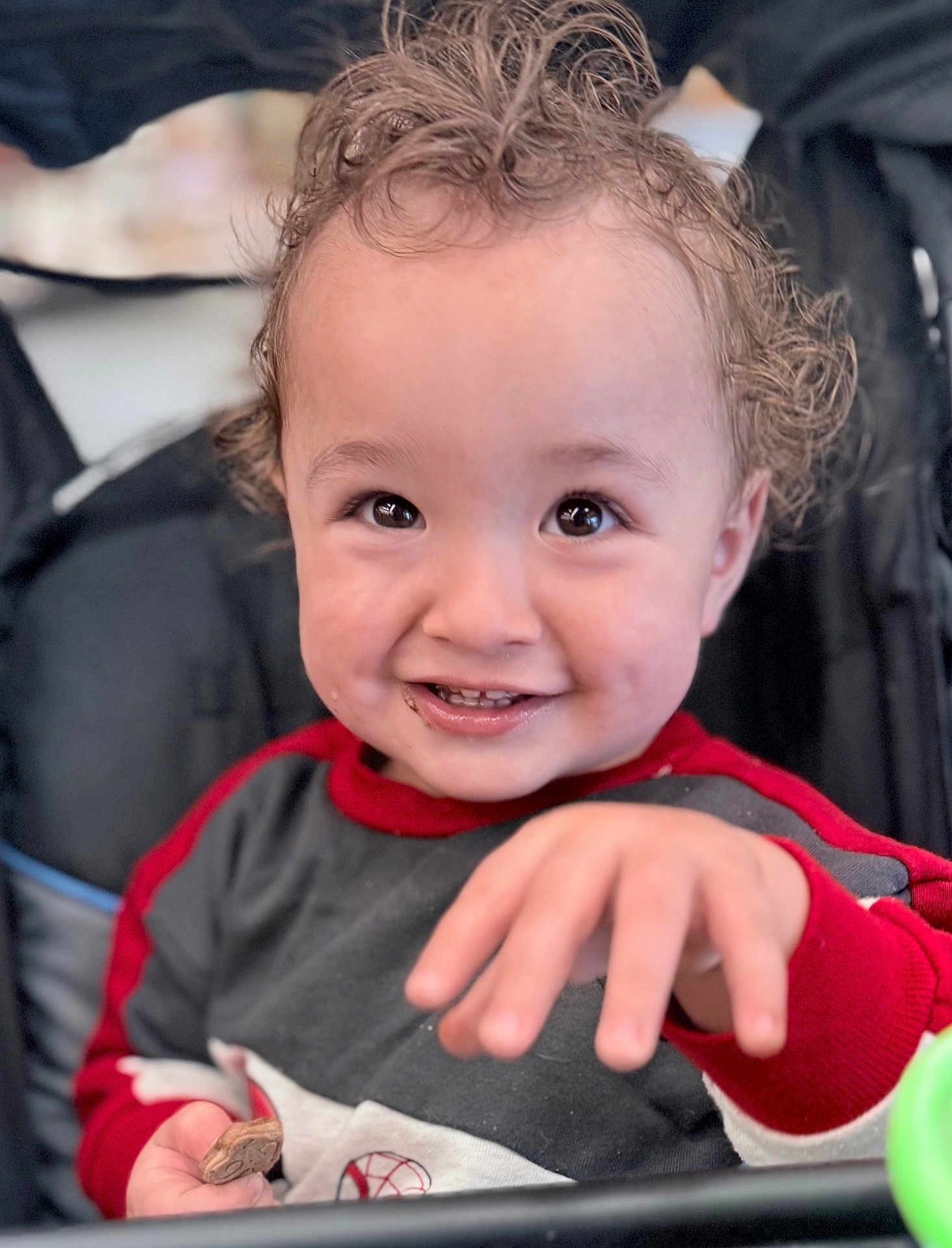 Luka is registered to the contest to win money with this photo: toddler, child, smiling, curly_hair, hand, cookie, clothing, red_sleeves, face, eyes, teeth, indoor, seat, stroller, happy, portrait, baby, person, cute, expression