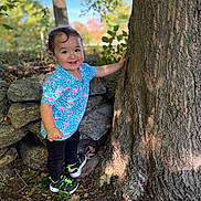 Luka is registered to the contest to win money with this photo: toddler, child, smiling, tree, outdoor, nature, rocks, leaves, forest, sunlight, blue_sky, shirt, black_pants, sneakers, happy, person, standing, young_child, daylight, casual_clothing