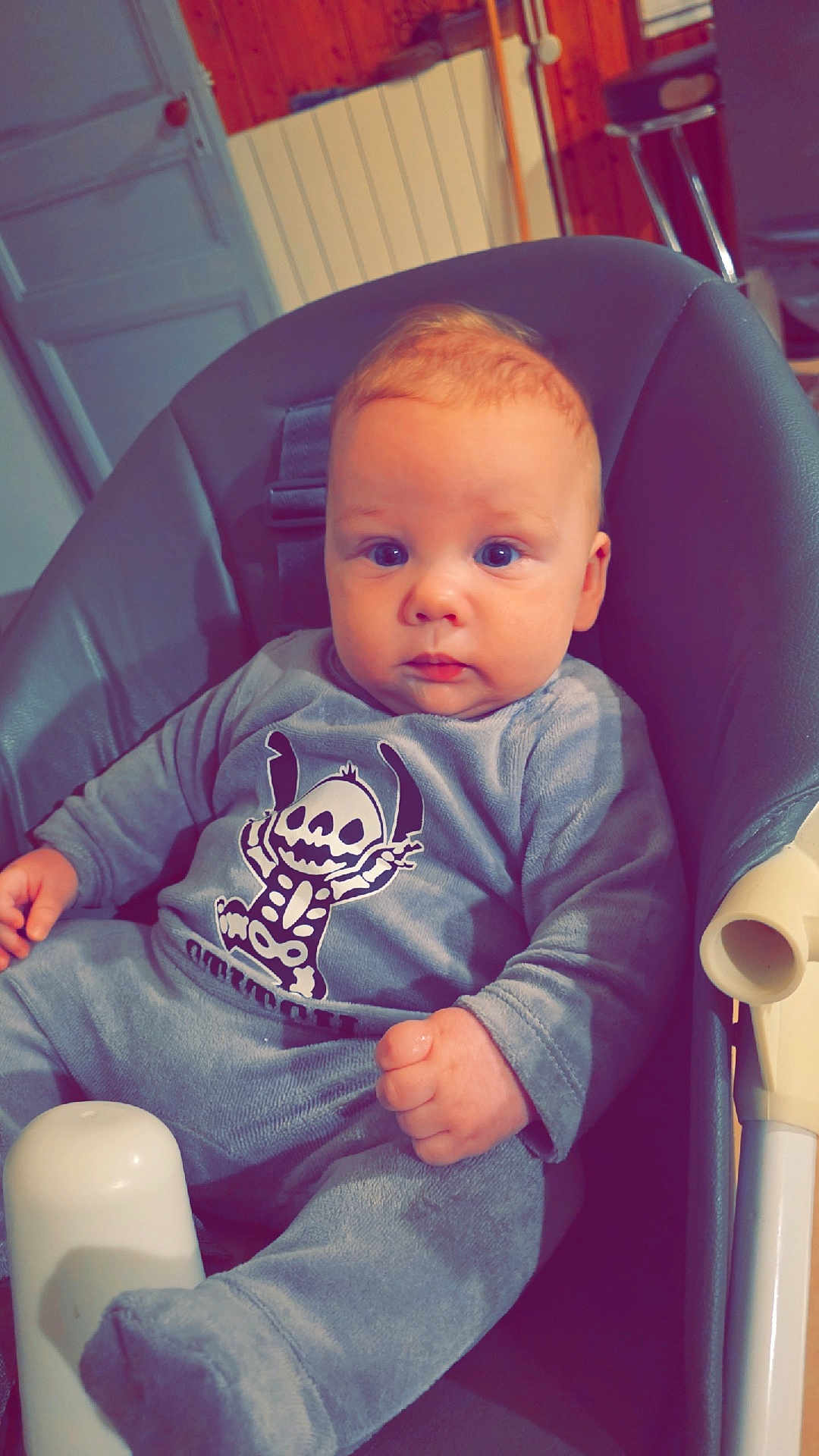 Raphaël participe au concours pour gagner de l'argent avec cette photo : baby, child, onesie, high_chair, blue_eyes, indoor, wooden_wall, cozy, infant, cute, portrait, skeleton_graphic, clothing, seat, furniture, young_child, face, hand, foot, expression