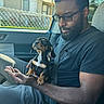 Scooby-Doo is registered to the contest to win money with this photo: man, glasses, black_tshirt, puppy, dog, car_interior, seatbelt, window, chain_link_fence, house, sunlight, beard, curious, lap, hand, casual_clothing, short_hair, indoor, portrait, pet