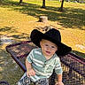 Rhett joined the competition — help win amazing prizes! baby, child, cowboy_hat, outdoor, park, picnic_table, seated, portrait, smiling, face, person, trees, grass, sunlight, shadows, nature, playful, striped_shirt, patterned_pants, stump