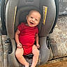 baby, camouflage, car_seat, child, comfortable, couch, face, floor, footwear, furniture, happy, indoor, infant, patterned, person, red_onesie, seat, shoe, smiling, wooden_floor