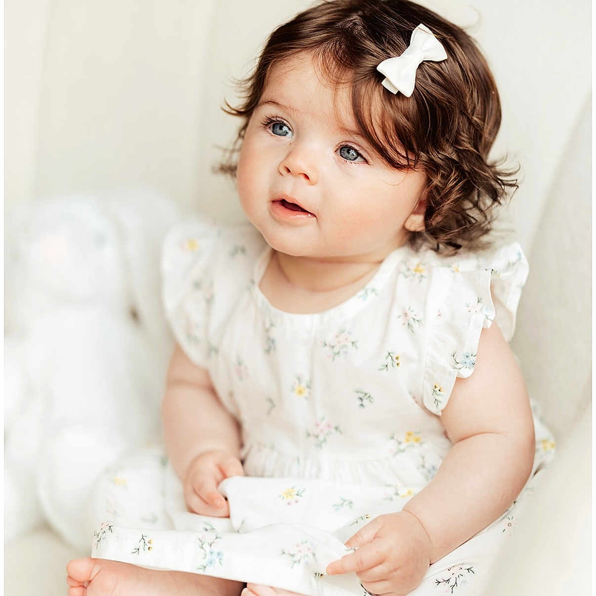 Lilah is registered to the contest to win money with this photo: adorable, baby, blue_eyes, child, couch, curly_hair, cute, expression, floral_dress, foot, hand, indoors, infant, person, portrait, sitting, skin, soft_light, white_bow, young