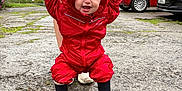 Arabelle joined the competition — help win amazing prizes! toddler, child, raincoat, red_clothing, boots, outdoor, walking, support, adult_hands, smiling, happy, pavement, car, house, cloudy_sky, person, hood, early_walking, joy, play