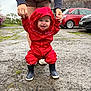 toddler, child, raincoat, red_clothing, boots, outdoor, walking, support, adult_hands, smiling, happy, pavement, car, house, cloudy_sky, person, hood, early_walking, joy, play