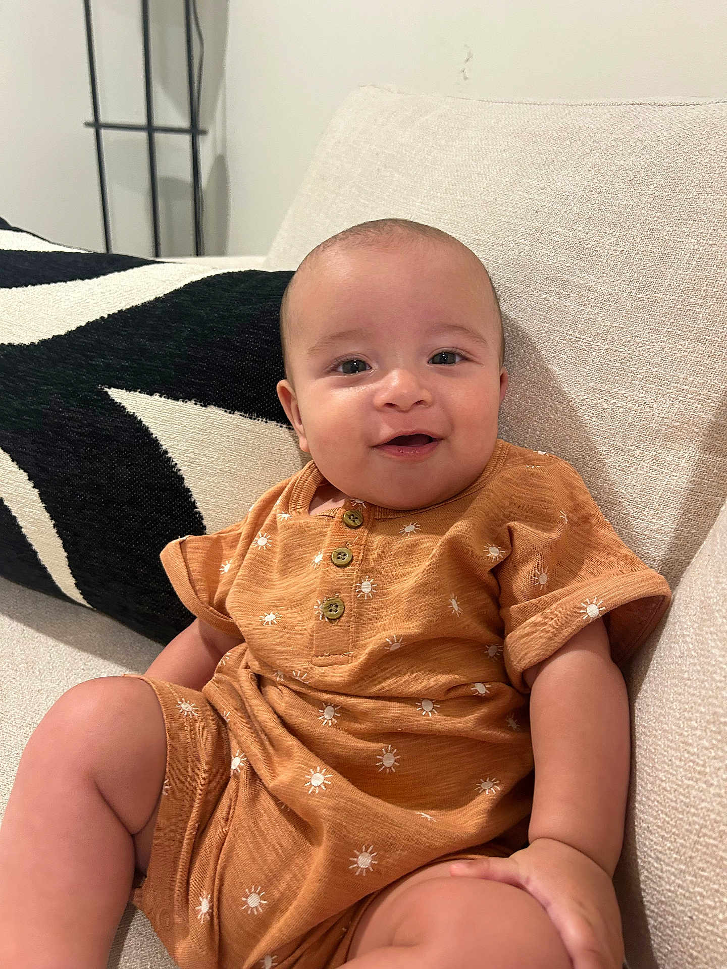 Osiah is registered to the contest to win money with this photo: baby, infant, smiling, orange_outfit, sun_pattern, couch, pillow, beige, black_and_white, happy, face, child, indoor, cute, portrait, sitting, comfortable, young, skin, head