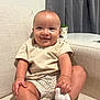 baby, child, smiling, couch, indoor, clothing, socks, shorts, shirt, face, hand, legs, furniture, curtain, plant, table, living_room, happy, sitting, person
