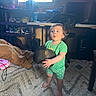toddler, child, barefoot, green_clothing, boot, carpet, indoor, furniture, desk, chair, shelf, shoes, window, television, expression, person, room, floor, toy, lighting