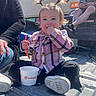 Sunshyne is registered to the contest to win money with this photo: toddler, child, outdoor, sunny, truck_bed, food, spoon, pigtails, plaid_shirt, sneakers, person, adult, casual_clothing, hand, container, blue_sky, building, signage, seated, relaxed