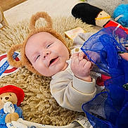 Henry joined the competition — help win amazing prizes! baby, smiling, bear_ears, fluffy_rug, toy, stuffed_animal, blue_fabric, hand_holding, indoor, child, cute, happy, infant, blanket, playtime, colorful, soft_texture, person, face, lying_down