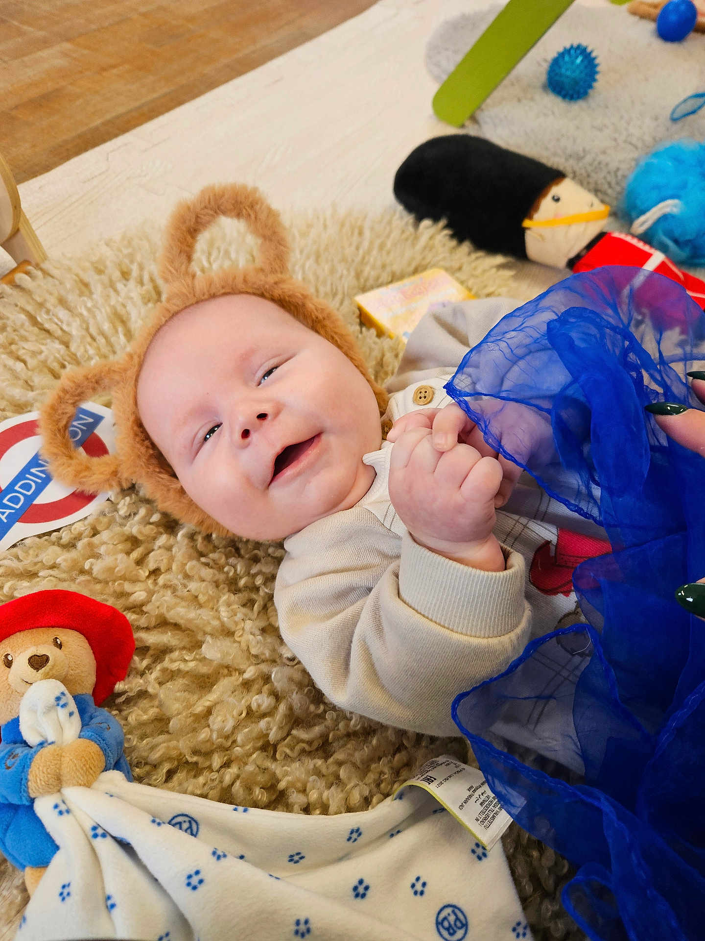 Henry joined the competition — help win amazing prizes! baby, smiling, bear_ears, fluffy_rug, toy, stuffed_animal, blue_fabric, hand_holding, indoor, child, cute, happy, infant, blanket, playtime, colorful, soft_texture, person, face, lying_down