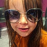 bangs, brown_hair, candid, child, close_up, face, fashion, indoor, kid, long_hair, mouth, nose, orange_shirt, playful, portrait, reflection_in_glasses, seat, smile, sunglasses, wall
